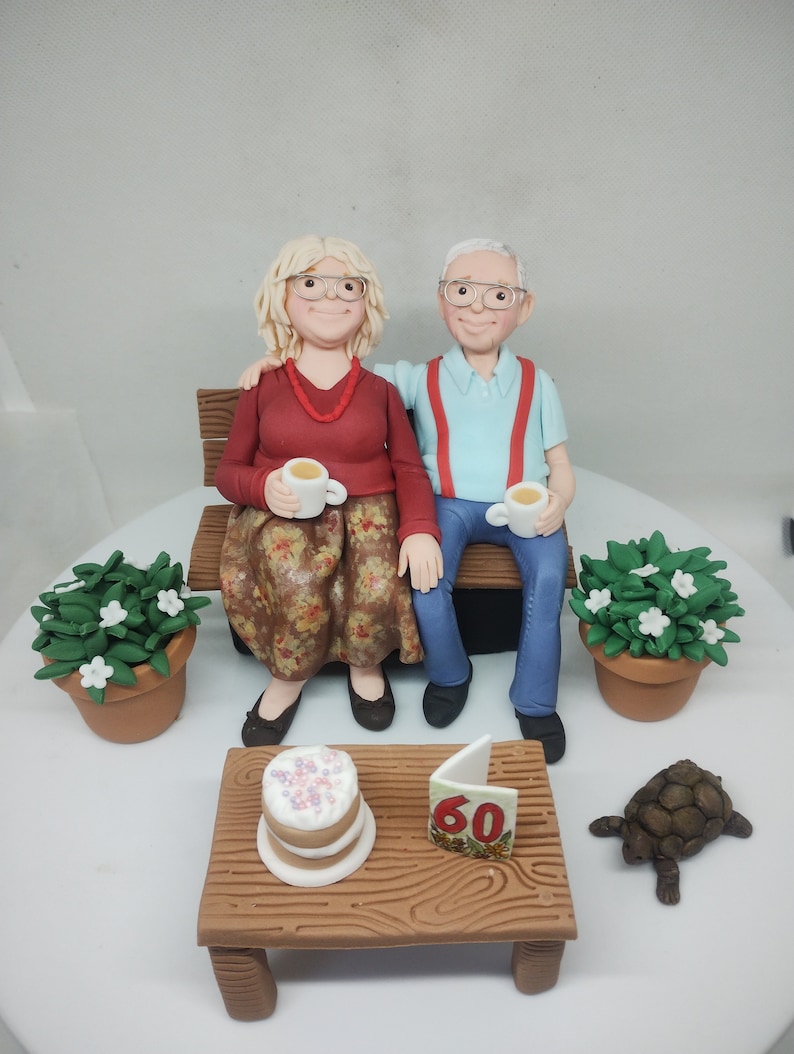 May include: A 60th anniversary cake with a couple sitting on a bench, holding cups of tea. The cake is decorated with a small cake, a card with the number 60, and a small turtle.