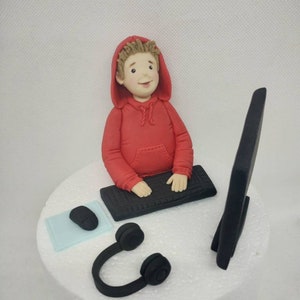 May include: A red hooded figure sits at a computer desk with a black keyboard, mouse, and headset. The figure is made of fondant and is a cake topper.