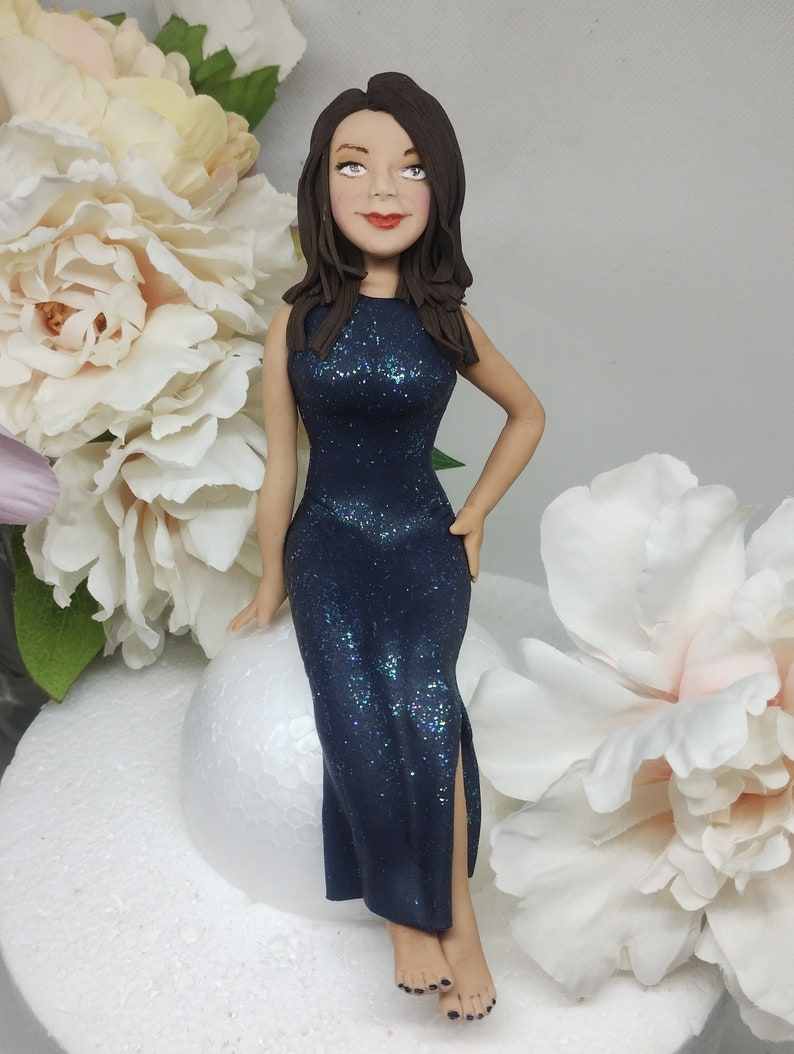 May include: A dark blue sparkly dress with a slit up the leg, worn by a woman with long brown hair and a neutral expression. She is sitting on a white round surface.