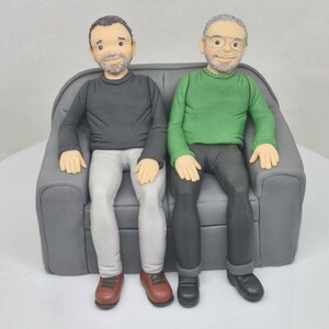 May include: Two figures of people sitting on a gray couch. The person on the left is wearing a black sweater, white pants, and brown shoes. The person on the right is wearing a green sweater, black pants, and black shoes.