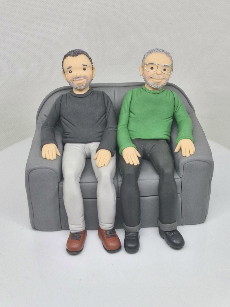 May include: Two figures of people sitting on a gray couch. The person on the left is wearing a black sweater, white pants, and brown shoes. The person on the right is wearing a green sweater, black pants, and black shoes.