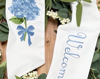 May include: Two white fabric ribbons featuring embroidery. One ribbon displays a blue hydrangea flower with a green stem and a blue bow. The other ribbon features the word Welcome embroidered in blue script lettering on the white fabric surface.