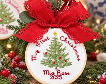 May include: A round wooden embroidery hoop ornament featuring white fabric with an embroidered green Christmas tree, red and pink ornaments, and a yellow star. The text reads My First Christmas, Mia Rose, and 2025. A large red lace-trimmed ribbon bow sits on top.