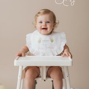 May include: A white ruffled bib featuring an embroidered birthday cake with pink and green balloons and the text Happy Birthday. The bib is worn by a child sitting in a white high chair. The image includes the text Twisted Magnolia by Amy.