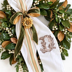May include: A round wreath featuring dark green and bronze magnolia leaves interspersed with clusters of small white berries. A white fabric ribbon with a striped pattern and a dark green velvet bow is centered with a gold flower.
