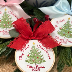 May include: Three round embroidery hoop ornaments featuring white fabric with a green embroidered Christmas tree, a yellow star, and red text reading My First Christmas and names like Mia Rose, topped with decorative pink, blue, or red fabric bows.