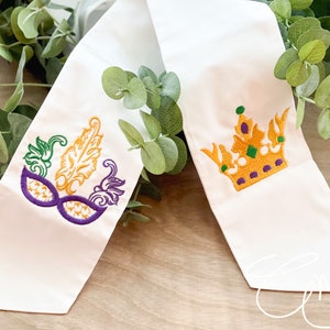 May include: A white fabric sash featuring two distinct embroidered designs. One end displays a purple and gold masquerade mask with green accents, while the other end features a gold crown with purple and green details.