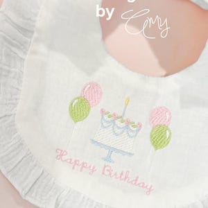 May include: A white ruffled baby bib featuring embroidered text that reads Twisted Magnolia by Cmy. The center design includes a birthday cake with a single candle, flanked by pairs of pink and green balloons, with the words Happy Birthday below.