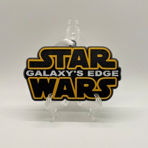 May include: Black and gold Star Wars Galaxy's Edge ornament. The ornament has a black background with gold lettering and a white ribbon for hanging. The ornament is displayed on a clear plastic stand.