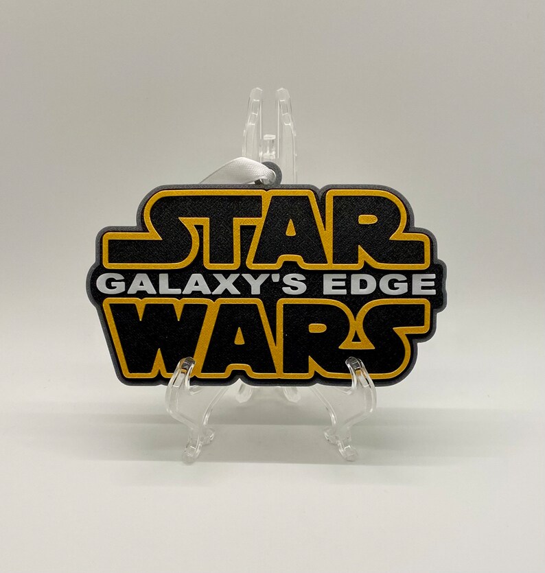 May include: Black and gold Star Wars Galaxy's Edge ornament. The ornament has a black background with gold lettering and a white ribbon for hanging. The ornament is displayed on a clear plastic stand.