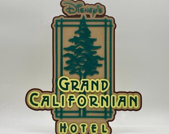 May include: A layered sign featuring the words "Disney's Grand Californian Hotel." The sign has a tan background with a green tree and a brown border. The text is in yellow and green.