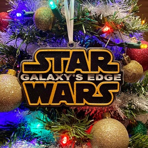 May include: A black and gold Star Wars Galaxy's Edge Christmas ornament. The ornament is rectangular with the words "STAR WARS" in gold and black. The ornament is hanging on a Christmas tree with colorful lights and gold and red ornaments.
