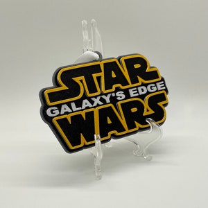 May include: A black and gold Star Wars Galaxy's Edge sign. The sign is in the shape of the Star Wars logo, with the words "STAR", "GALAXY'S EDGE", and "WARS" in gold and white lettering. The sign is displayed on a clear plastic stand.