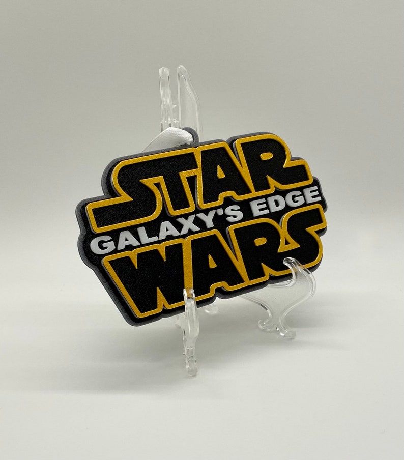 May include: A black and gold Star Wars Galaxy's Edge sign. The sign is in the shape of the Star Wars logo, with the words "STAR", "GALAXY'S EDGE", and "WARS" in gold and white lettering. The sign is displayed on a clear plastic stand.