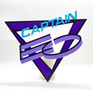 Puede incluir: Triángulo morado y negro con un diseño estilizado morado y negro en su interior. La palabra "CAPTAIN" está escrita en letras azules en la parte superior del triángulo.