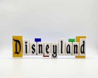 May include: A white sign with the word "Disneyland" spelled out in black letters. Each letter is on a separate block, with the "D" on a yellow block.