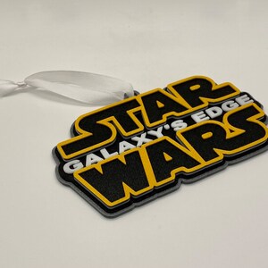 May include: A layered ornament featuring the text "STAR WARS GALAXY'S EDGE" in black, yellow, and gray. The ornament has a white ribbon for hanging. The design is a 3D rendering of the Star Wars logo.