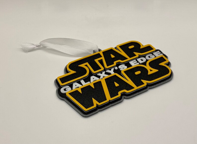 May include: A layered ornament featuring the text "STAR WARS GALAXY'S EDGE" in black, yellow, and gray. The ornament has a white ribbon for hanging. The design is a 3D rendering of the Star Wars logo.