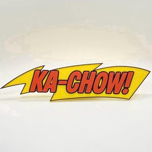 May include: A yellow lightning bolt-shaped graphic with the words "KA-CHOW!" in red, outlined in black. The design is reminiscent of a comic book or pop art style, with a bold and energetic aesthetic.