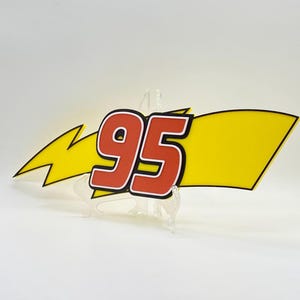 Puede incluir: Un letrero decorativo con el n&uacute;mero "95" en rojo con un contorno blanco, sobre un fondo amarillo con forma de pancarta y un rayo amarillo. El letrero est&aacute; sostenido por un soporte transparente.