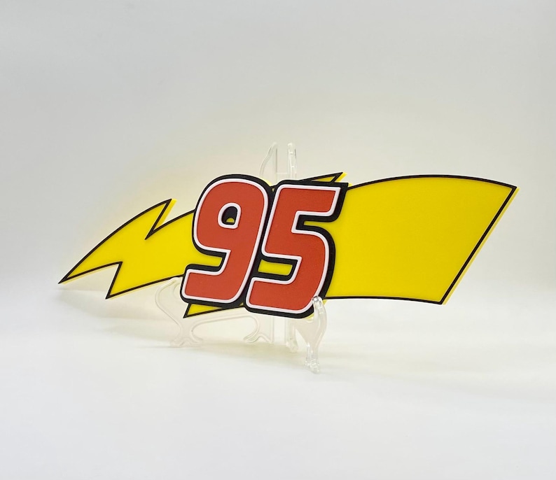 Puede incluir: Un letrero decorativo con el n&uacute;mero "95" en rojo con un contorno blanco, sobre un fondo amarillo con forma de pancarta y un rayo amarillo. El letrero est&aacute; sostenido por un soporte transparente.