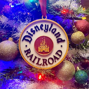 May include: A white and gold Christmas ornament with the text "Disneyland Railroad" and a red image of the Sleeping Beauty Castle.