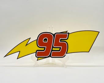 Puede incluir: Un letrero decorativo con el número "95" en rojo con contorno blanco, sobre un rayo amarillo y una bandera amarilla. El diseño es audaz y gráfico, adecuado para decoración temática.