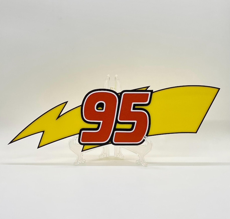 Puede incluir: Un letrero decorativo con el n&uacute;mero "95" en rojo con contorno blanco, sobre un rayo amarillo y una bandera amarilla. El dise&ntilde;o es audaz y gr&aacute;fico, adecuado para decoraci&oacute;n tem&aacute;tica.