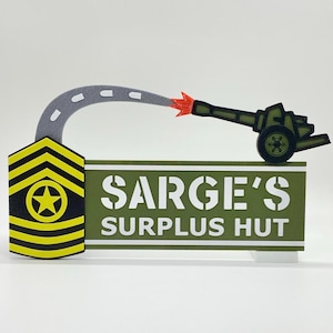 Può includere: Un cartello decorativo con la scritta "SARGE'S SURPLUS HUT". Il cartello presenta un tema militare con un gallone giallo e nero, una strada grigia e un cannone verde che spara un proiettile arancione.