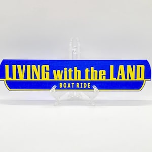 Può includere: Un cartello blu con testo giallo che recita "LIVING with the LAND BOAT RIDE". Il cartello ha un bordo giallo ed è esposto su un supporto trasparente. Il cartello è rettangolare con bordi curvi.