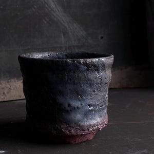 Könnte beinhalten: Ein zylindrischer Keramikbecher mit rauer, unebener Textur. Der obere Teil ist mit einer dunklen, gesprenkelten Glasur in Anthrazit, Schwarz und bläulichen Nuancen versehen, während der Boden aus unglasiertem, dunkelrotbraunem Ton besteht.
