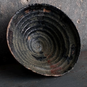 Könnte beinhalten: Eine tiefe, handgefertigte Keramikschale mit einer dunklen, gesprenkelten Glasur in Schwarz und Anthrazit. Das Innere weist ausgeprägte, konzentrische Spiralrillen auf, die vom Zentrum ausgehen, durchsetzt mit erdigen Braun- und Rosttönen.