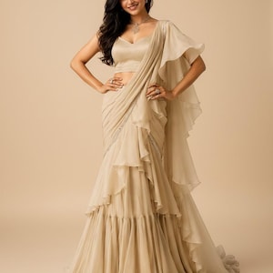 May include: A champagne-colored saree-style gown featuring a sleeveless bodice with a sweetheart neckline, a draped skirt with cascading ruffles, and a matching shoulder drape. The garment is crafted from a lightweight, flowing fabric.