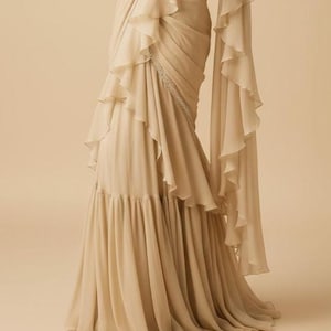 May include: A floor-length beige saree featuring cascading ruffles along the drape and a pleated lower section. The garment includes a thin, embellished border with silver-toned detailing that runs diagonally across the front of the fabric.