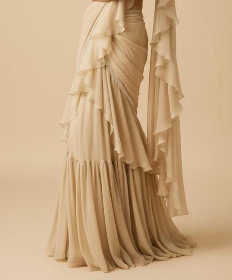 May include: A floor-length beige saree featuring cascading ruffles along the drape and a pleated lower section. The garment includes a thin, embellished border with silver-toned detailing that runs diagonally across the front of the fabric.