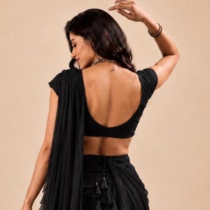 Puede incluir: Una blusa negra de manga corta con escote redondo profundo, combinada con una falda negra a juego que presenta un dise&ntilde;o de tela drapeada y superpuesta, con un cierre de lazo en la cintura y peque&ntilde;as borlas negras colgando en el centro.