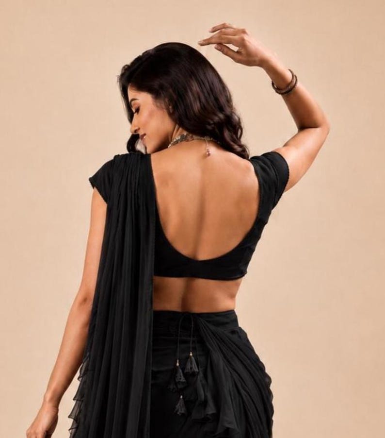 Puede incluir: Una blusa negra de manga corta con escote redondo profundo, combinada con una falda negra a juego que presenta un dise&ntilde;o de tela drapeada y superpuesta, con un cierre de lazo en la cintura y peque&ntilde;as borlas negras colgando en el centro.