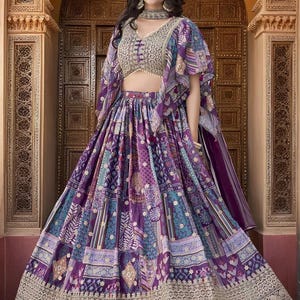 May include: A vibrant purple and blue patterned lehenga, a traditional Indian garment, featuring a detailed embroidered bodice and a matching shawl. The skirt has a patchwork design with floral and geometric motifs, and a gold embroidered border.