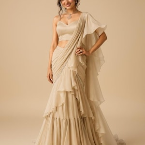 May include: A beige sleeveless blouse with a sweetheart neckline paired with a matching floor-length skirt featuring layered ruffles and a draped sash detail that cascades from the shoulder down to the hemline in a flowing silhouette.