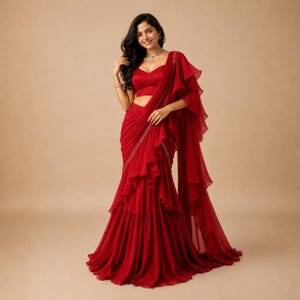 Puede incluir: Un sari de color rojo vibrante que presenta una blusa sin mangas con escote corazón y una falda larga hasta el suelo con capas de volantes en cascada. La prenda está confeccionada en un tejido ligero y semitransparente con un pallu fluido.