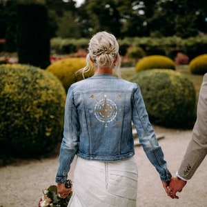Könnte beinhalten: Ein Paar hält sich an den Händen, die Frau trägt ein weißes Kleid und eine Jeansjacke mit einem goldenen Kompass-Design auf dem Rücken. Der Mann trägt eine braune Anzugsjacke.