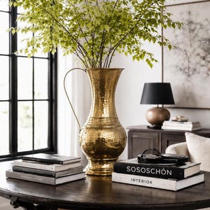Könnte beinhalten: Eine goldfarbene, gehämmerten Metallvase mit Henkel, gefüllt mit grünen, belaubten Zweigen, steht auf einem dunklen Holztisch. Bücher und eine schwarze Lampe befinden sich ebenfalls auf dem Tisch. Die Vase ist ein markantes Dekorationselement.