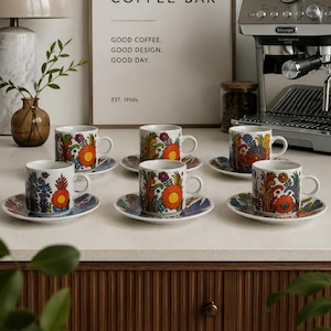 Könnte beinhalten: Ein Set aus sechs weißen Espresso-Tassen aus Keramik mit passenden Untertassen, die jeweils mit einem lebendigen, mehrfarbigen Blumenmuster in Orange, Blau und Grün verziert sind. Die Tassen stehen in zwei Reihen auf einer hellen Arbeitsplatte.