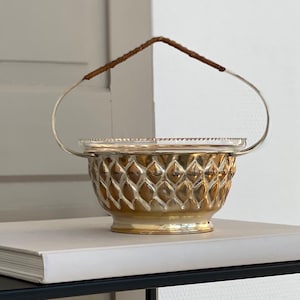 Könnte beinhalten: Eine goldfarbene Zierschale mit Rautenmuster, silbernem Griff und braunem Ledergriff. Die Schale steht auf einem weißen Buch, das sich auf einem schwarzen Tisch befindet.