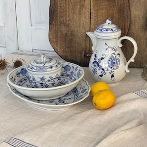 Può includere: Un set di brocca per la panna e zuccheriera in porcellana bianca con motivi floreali blu, un brocca in porcellana bianca con motivi floreali blu e limoni su una tovaglia di lino bianca con strisce blu. Il set è su un tavolo di legno con due taglieri di legno sullo sfondo.