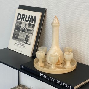 Könnte beinhalten: Ein Set aus fünf weißen Alabaster-Schnapsgläsern und einer passenden Karaffe auf einem schwarzen Regal mit einem Buch mit dem Titel "Paris Vu Du Ciel" und anderen Büchern mit den Titeln "Icon" und "Drum".