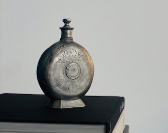 Könnte beinhalten: Eine runde, dekorative Flasche aus Silber mit einem Stopfen und gravierten Details, darunter das Jahr 1895, die auf einem Stapel Bücher steht.