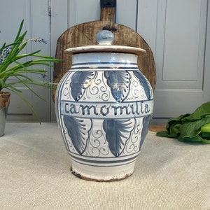 Può includere: Un vaso di ceramica bianca con un disegno floreale blu e bianco e la parola "camomilla" scritta su di esso. Il vaso ha un coperchio ed è seduto su una tovaglia bianca.