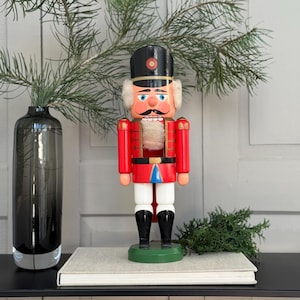 May include: A wooden nutcracker soldier in a red uniform with a black hat and white hair. The nutcracker stands on a green base and is displayed on a shelf with a vase and greenery.
