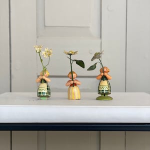 Könnte beinhalten: Drei Holzfiguren von Mädchen, die Blumen halten. Jedes Mädchen trägt ein Kleid in einer anderen Farbe und hält eine andere Blume. Die Blumen sind weiß und grün.